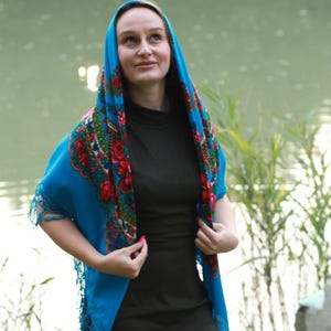 May include: A woman wearing a teal headscarf with a floral pattern of red roses and green leaves. The scarf is draped over the head and shoulders. She is wearing a dark green dress. The background is a lake.