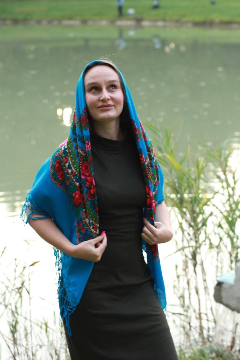 May include: A woman wearing a teal headscarf with a floral pattern of red roses and green leaves. The scarf is draped over the head and shoulders. She is wearing a dark green dress. The background is a lake.