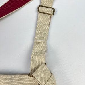Puede incluir: Una bolsa de lona beige con una correa de hombro ajustable de color rojo y beige. La correa tiene una hebilla de lat&oacute;n.