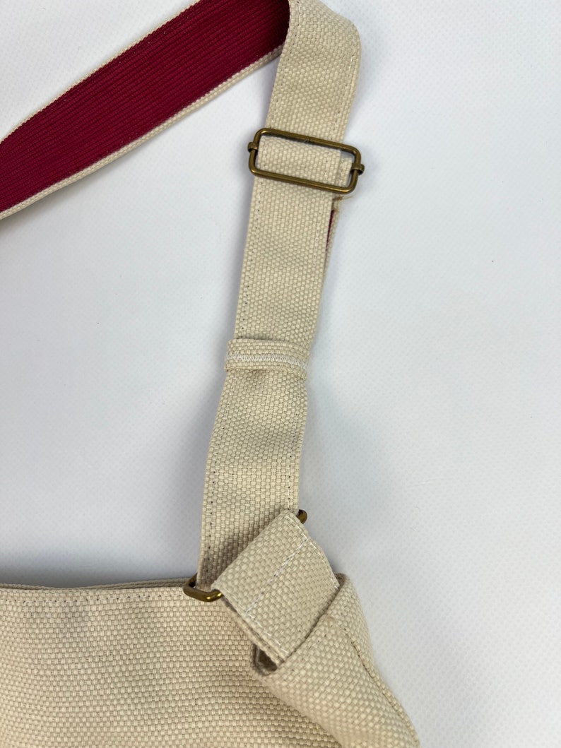 Puede incluir: Una bolsa de lona beige con una correa de hombro ajustable de color rojo y beige. La correa tiene una hebilla de lat&oacute;n.