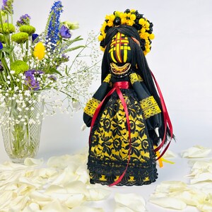 K&ouml;nnte beinhalten: Handgefertigte Puppe mit einem schwarzen und goldenen Kleid, einem gelben Blumenkranz und langen schwarzen Haaren. Die Puppe hat ein bemaltes Gesicht mit roten und gelben Akzenten. Rote B&auml;nder h&auml;ngen von der Taille. Wei&szlig;e Bl&uuml;tenbl&auml;tter umgeben die Puppe.