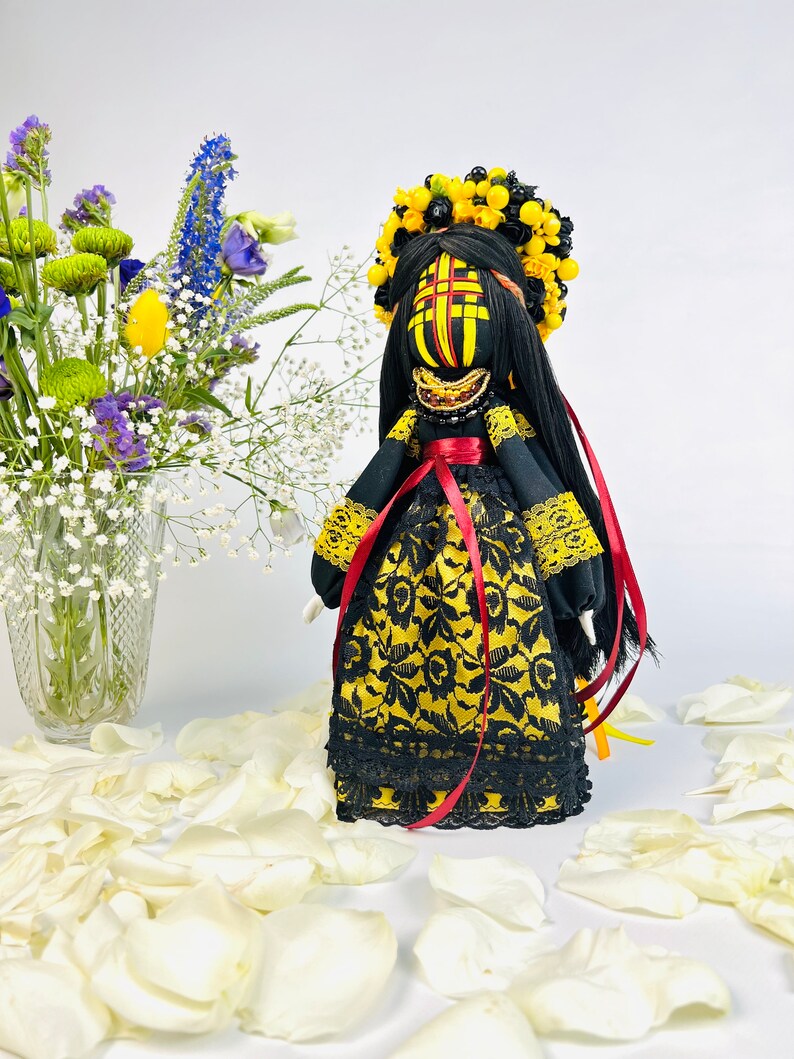 K&ouml;nnte beinhalten: Handgefertigte Puppe mit einem schwarzen und goldenen Kleid, einem gelben Blumenkranz und langen schwarzen Haaren. Die Puppe hat ein bemaltes Gesicht mit roten und gelben Akzenten. Rote B&auml;nder h&auml;ngen von der Taille. Wei&szlig;e Bl&uuml;tenbl&auml;tter umgeben die Puppe.