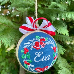 K&ouml;nnte beinhalten: Ein blauer Weihnachtsbaumschmuck mit einem rot-wei&szlig;en Band. Der Schmuck ist mit einem Kranz aus Stechpalme, Glocken und dem Namen "Eva" verziert.