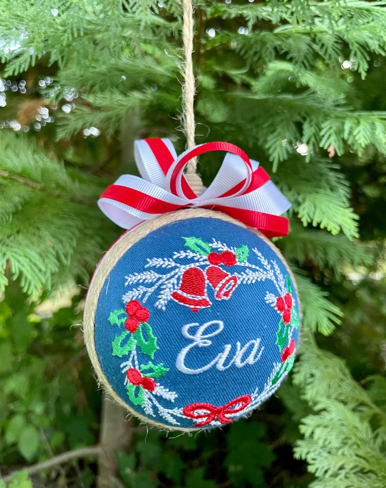 K&ouml;nnte beinhalten: Ein blauer Weihnachtsbaumschmuck mit einem rot-wei&szlig;en Band. Der Schmuck ist mit einem Kranz aus Stechpalme, Glocken und dem Namen "Eva" verziert.