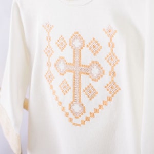 May include: White t-shirt with a gold embroidered cross design.