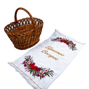 Pu&ograve; includere: Un asciugamano da cucina bianco ricamato con un motivo floreale e il testo "Buona Pasqua" in lettere dorate. Nell'immagine &egrave; presente anche un cesto di vimini marrone.