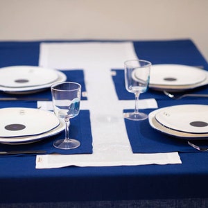 May include: A table setting with four place settings. Each setting includes a white plate with a black center, a glass, a fork, and a knife. The table is covered with a blue tablecloth and white runner.