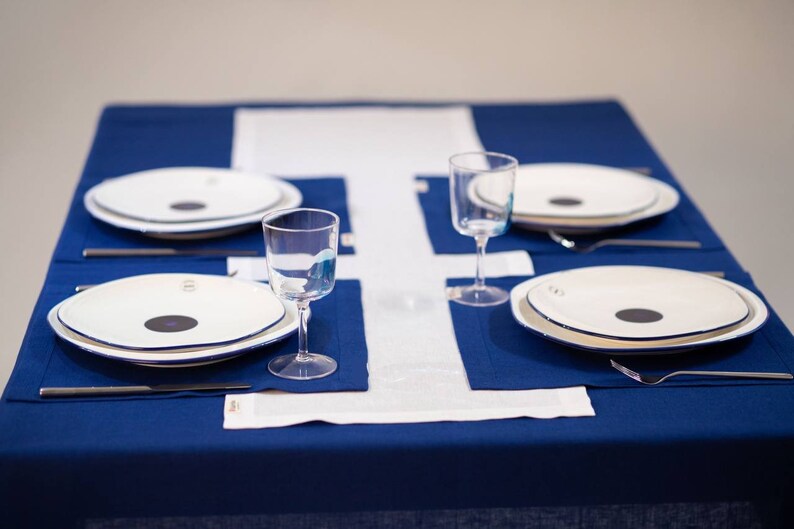 May include: A table setting with four place settings. Each setting includes a white plate with a black center, a glass, a fork, and a knife. The table is covered with a blue tablecloth and white runner.