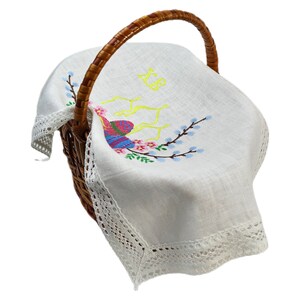 May include: A wicker basket with a brown handle, containing a white linen cloth. The cloth is embroidered with a colourful bird, pink flowers, and blue branches. The cloth has a delicate lace trim and yellow embroidered initials.