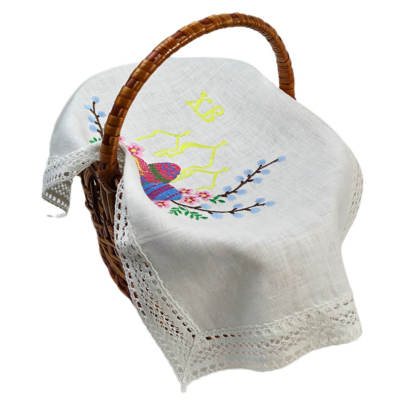 May include: A wicker basket with a brown handle, containing a white linen cloth. The cloth is embroidered with a colourful bird, pink flowers, and blue branches. The cloth has a delicate lace trim and yellow embroidered initials.