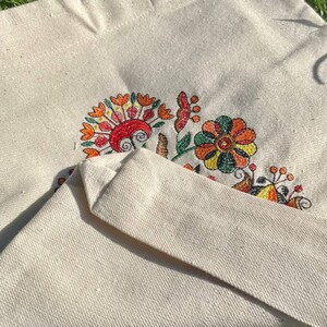 May include: A white canvas tote bag with a colorful embroidered floral design. The flowers are in shades of orange, yellow, green, and red.