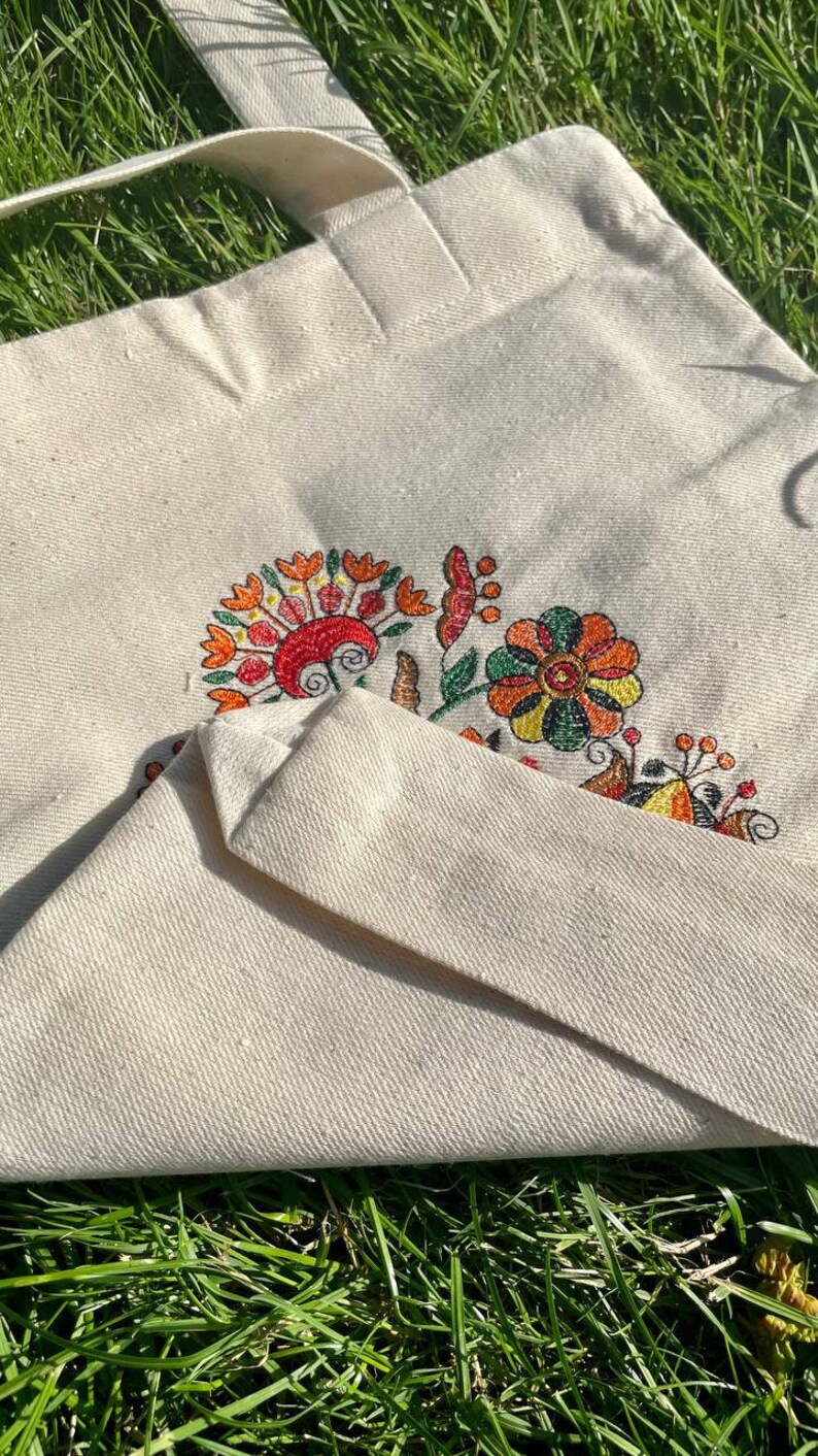 May include: A white canvas tote bag with a colorful embroidered floral design. The flowers are in shades of orange, yellow, green, and red.
