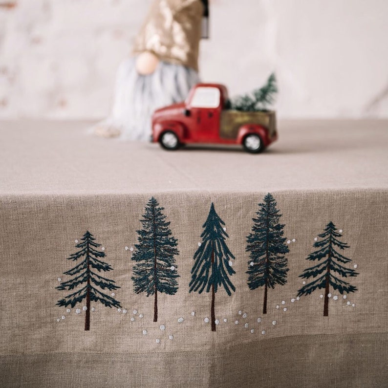 May include: A beige tablecloth with an embroidered forest scene featuring five dark green pine trees with brown trunks and white accents. A red toy truck with a Christmas tree in the bed is in the background.