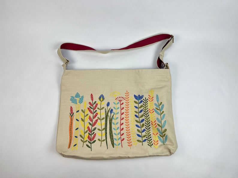 Puede incluir: Una bolsa de lona beige con una correa de hombro roja y un dise&ntilde;o floral bordado en tonos de azul, amarillo, verde y rojo.