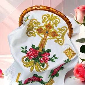 May include: White fabric with a cross embroidered in gold and red beads. The cross is surrounded by pink roses and green leaves. The fabric is in a wicker basket.