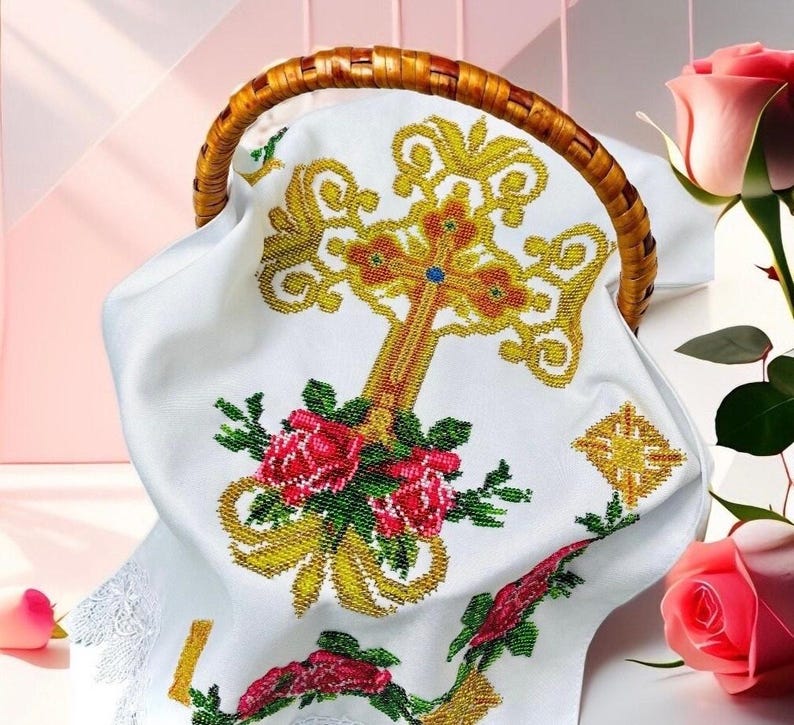 May include: White fabric with a cross embroidered in gold and red beads. The cross is surrounded by pink roses and green leaves. The fabric is in a wicker basket.