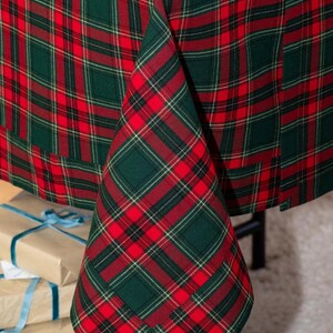 May include: A festive red and green plaid tablecloth. The fabric features a classic tartan pattern, perfect for holiday gatherings. The tablecloth is draped over a table, with a corner folded to show the pattern. Presents are visible in the background.