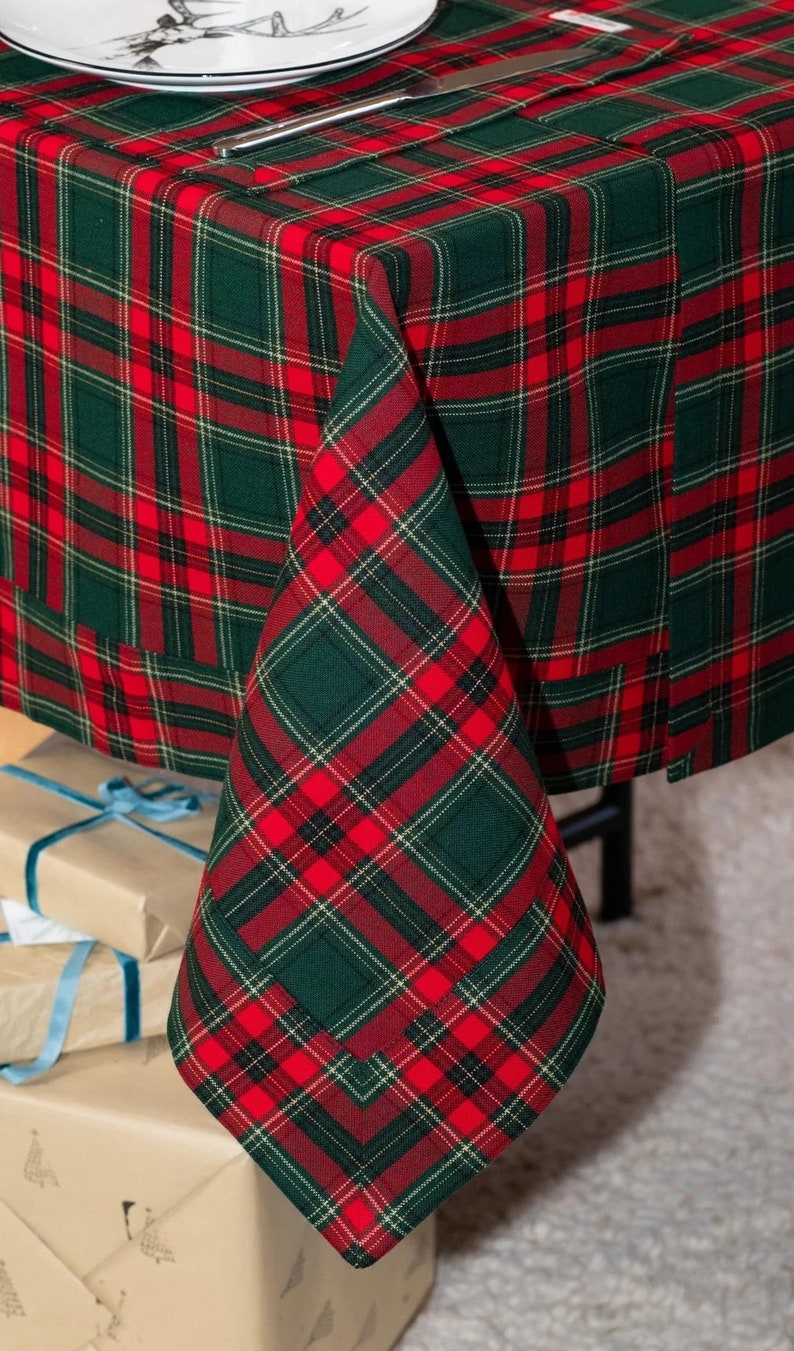 May include: A festive red and green plaid tablecloth. The fabric features a classic tartan pattern, perfect for holiday gatherings. The tablecloth is draped over a table, with a corner folded to show the pattern. Presents are visible in the background.