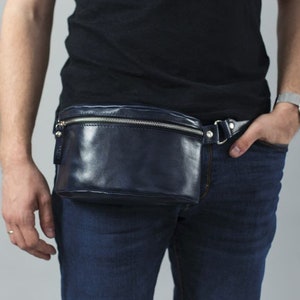 May include: A dark blue leather fanny pack with a silver zipper and buckle. The fanny pack is worn around the waist of a person wearing a black shirt and blue jeans.