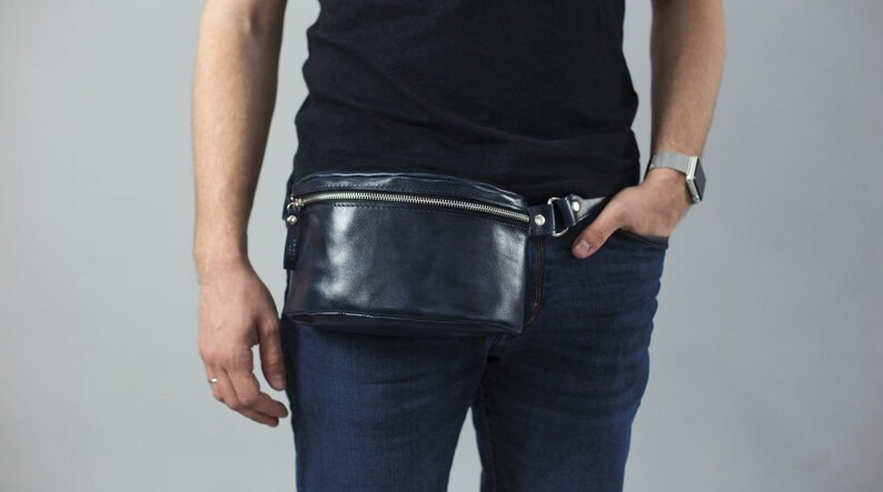 May include: A dark blue leather fanny pack with a silver zipper and buckle. The fanny pack is worn around the waist of a person wearing a black shirt and blue jeans.