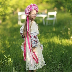 May include: A young girl wearing a white embroidered dress with a pink ribbon and a pink flower crown stands in a field of green grass.