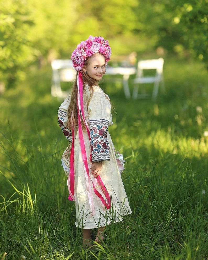 May include: A young girl wearing a white embroidered dress with a pink ribbon and a pink flower crown stands in a field of green grass.