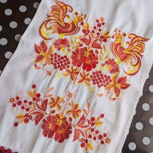 Peut inclure: Serviette en lin blanc avec broderie vibrante repr&eacute;sentant deux oiseaux stylis&eacute;s, des fleurs et des baies dans des tons de rouge, orange et jaune. Le bord inf&eacute;rieur est orn&eacute; d'une bordure en dentelle rouge. Textile d&eacute;coratif.