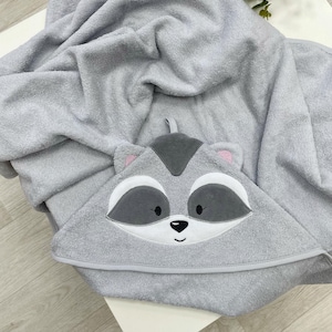 May include: A light grey hooded baby towel with a raccoon face design. The towel features a hood with a raccoon face, including grey and white details, and pink inner ears. The towel is made of soft, absorbent material.