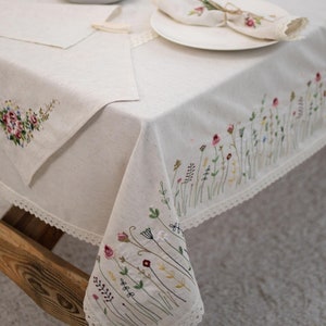 May include: A white linen tablecloth with delicate floral embroidery. The tablecloth features a lace trim and is decorated with a pattern of small flowers and leaves in shades of pink, green, and white.