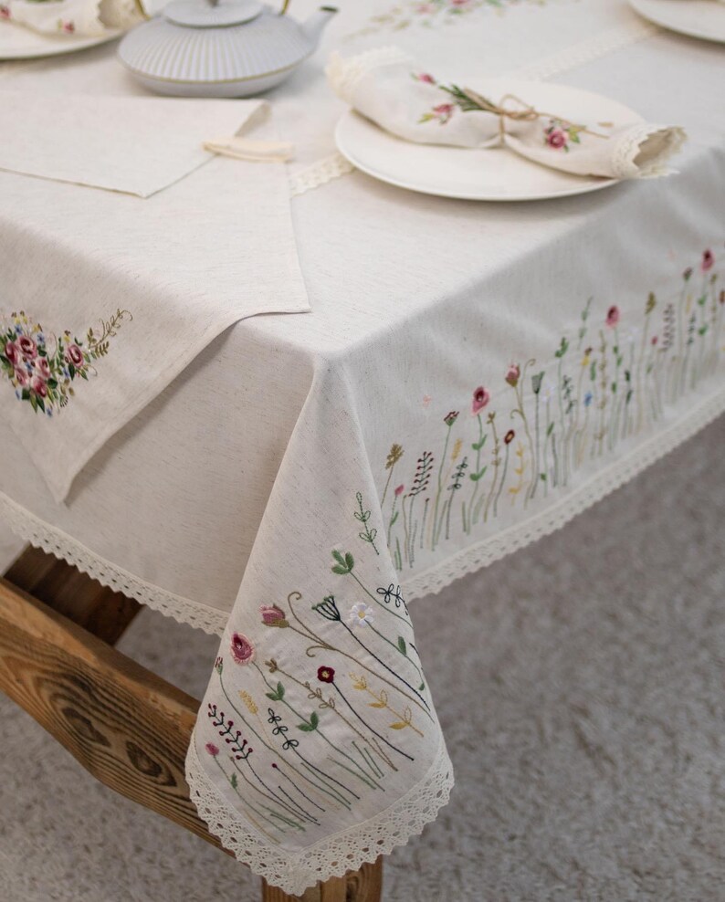 May include: A white linen tablecloth with delicate floral embroidery. The tablecloth features a lace trim and is decorated with a pattern of small flowers and leaves in shades of pink, green, and white.