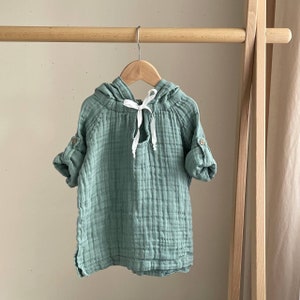 May include: A sage green hooded shirt with long sleeves and a white ribbon tie at the neck. The shirt is made of a soft, lightweight fabric and has a relaxed fit.