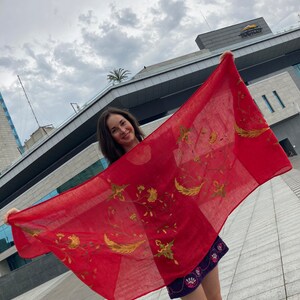 Puede incluir: Una bufanda roja con bordados dorados de flores y hojas. La bufanda est&aacute; siendo sostenida por una persona frente a un edificio.