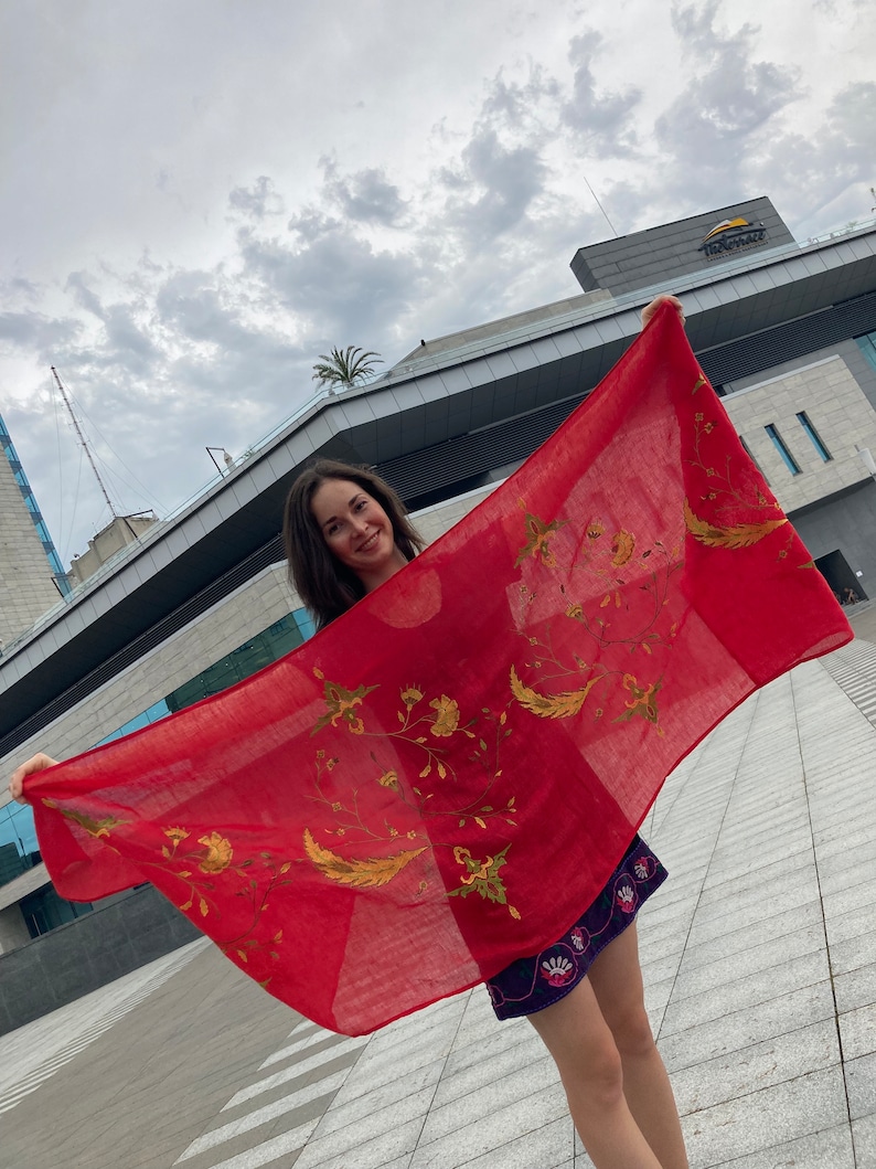 Puede incluir: Una bufanda roja con bordados dorados de flores y hojas. La bufanda est&aacute; siendo sostenida por una persona frente a un edificio.