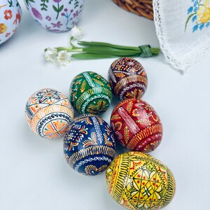 Pu&ograve; includere: Una collezione di uova di Pasqua decorate con colori vivaci, tra cui arancione, verde, rosso e giallo. Ogni uovo presenta intricati disegni dipinti a mano. Sono visibili anche due bicchieri a motivi floreali e un cestino di vimini, che suggeriscono un tema primaverile.