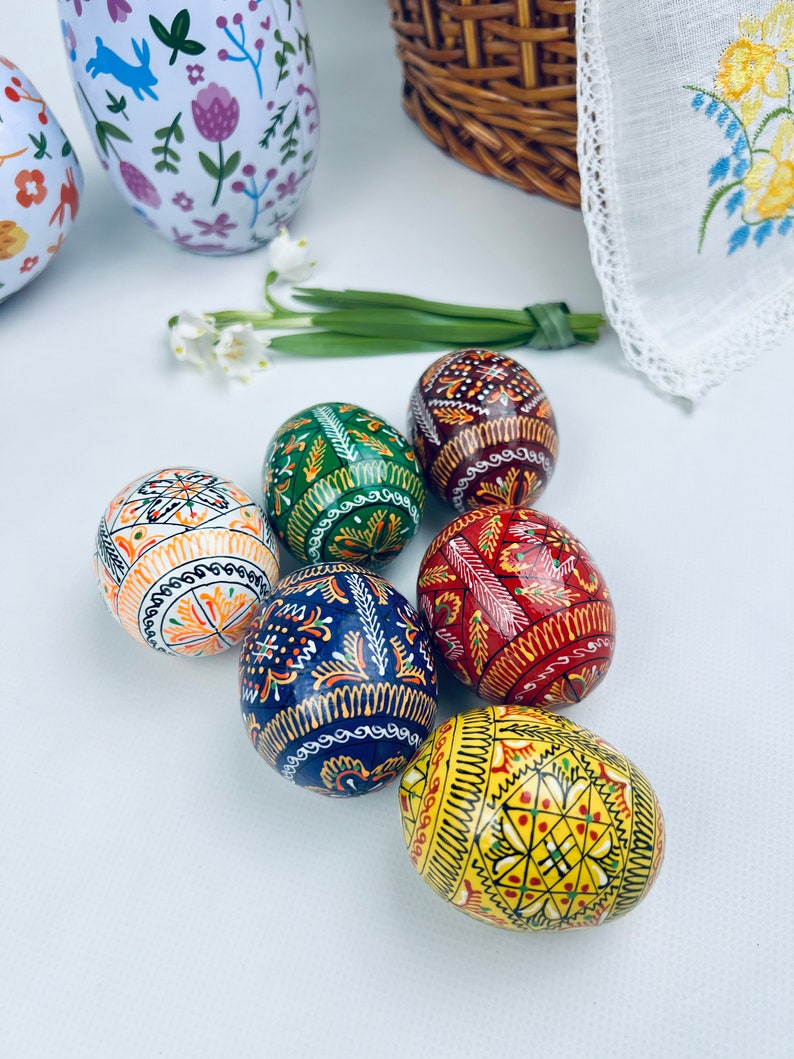 Pu&ograve; includere: Una collezione di uova di Pasqua decorate con colori vivaci, tra cui arancione, verde, rosso e giallo. Ogni uovo presenta intricati disegni dipinti a mano. Sono visibili anche due bicchieri a motivi floreali e un cestino di vimini, che suggeriscono un tema primaverile.