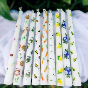 May include: A collection of white taper candles, each hand-painted with colorful designs. The designs include bees, carrots, lemons, blueberries, avocados, and other nature-inspired motifs. These decorative candles are perfect for adding a touch of whimsy to any space.