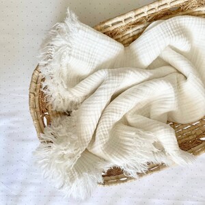 K&ouml;nnte beinhalten: Eine wei&szlig;e Musselin-Babydecke mit ausgefransten R&auml;ndern, gefaltet und in einen geflochtenen Korb gelegt.
