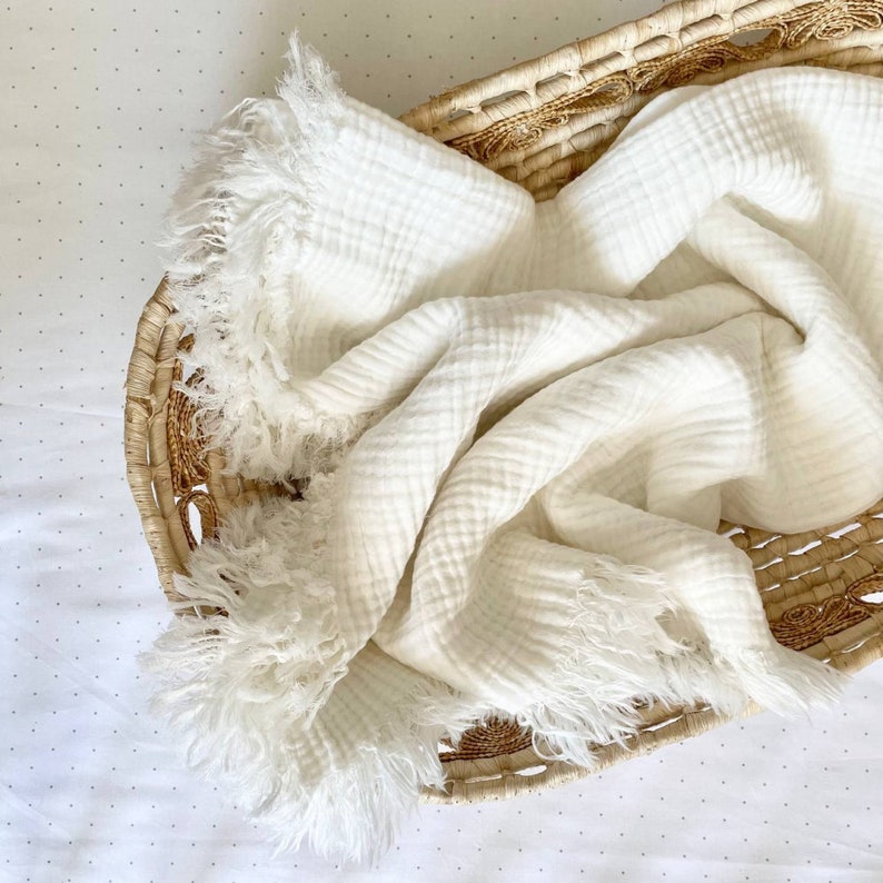 K&ouml;nnte beinhalten: Eine wei&szlig;e Musselin-Babydecke mit ausgefransten R&auml;ndern, gefaltet und in einen geflochtenen Korb gelegt.