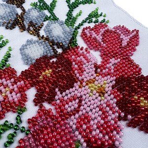 May include: Close-up of a beaded floral design on white fabric. The design features pink, red, and white flowers with green leaves.