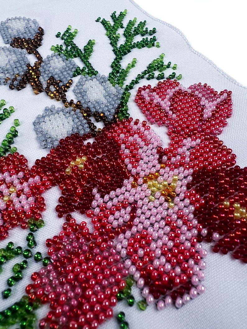 May include: Close-up of a beaded floral design on white fabric. The design features pink, red, and white flowers with green leaves.