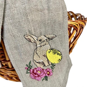 May include: A beige linen Easter napkin with an embroidered design of a bunny, a yellow egg with floral details, and pink flowers. The napkin is draped over a wicker basket.