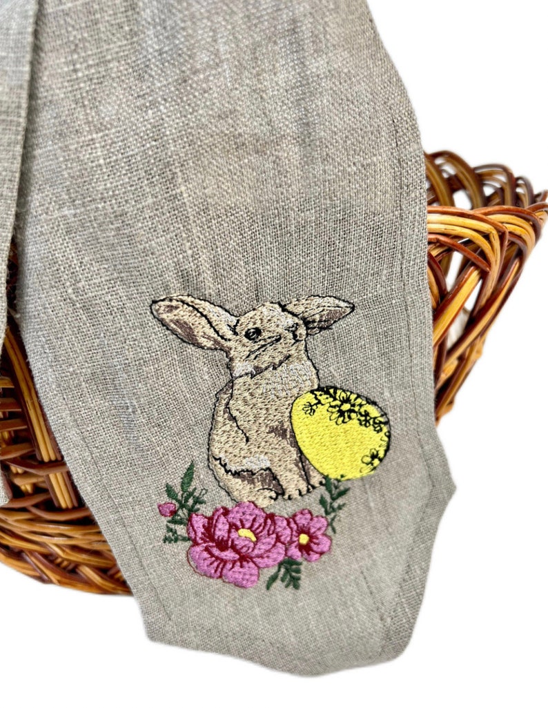 May include: A beige linen Easter napkin with an embroidered design of a bunny, a yellow egg with floral details, and pink flowers. The napkin is draped over a wicker basket.