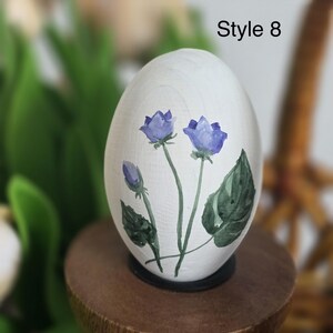 May include: A white, egg-shaped decorative item with hand-painted blue flowers and green leaves. The text "Style 8" is printed in black at the top. The item is displayed on a wooden stand.