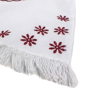 May include: White textile with a burgundy embroidered design. The design includes a circular pattern with deer and geometric shapes, and snowflake-like accents. The edge has a fringe.