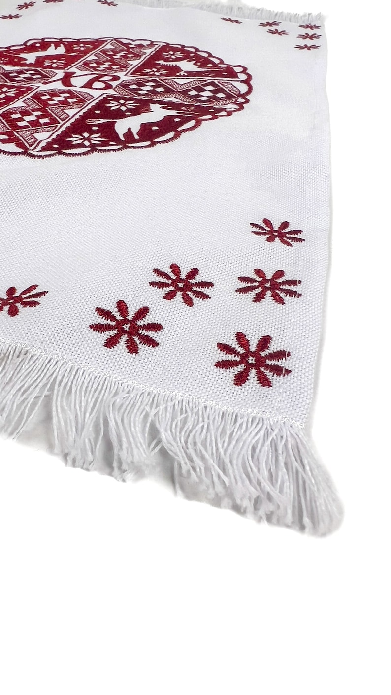 May include: White textile with a burgundy embroidered design. The design includes a circular pattern with deer and geometric shapes, and snowflake-like accents. The edge has a fringe.