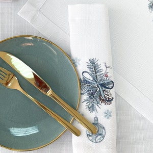 K&ouml;nnte beinhalten: Ein t&uuml;rkisfarbener Teller mit Goldrand, eine goldene Gabel und ein goldenes Messer liegen auf einer wei&szlig;en Tischdecke. Rechts befindet sich eine wei&szlig;e Serviette mit einem blau-silbernen Weihnachtsbaumdesign. Im Hintergrund ein wei&szlig;er Tischset mit passendem Design.