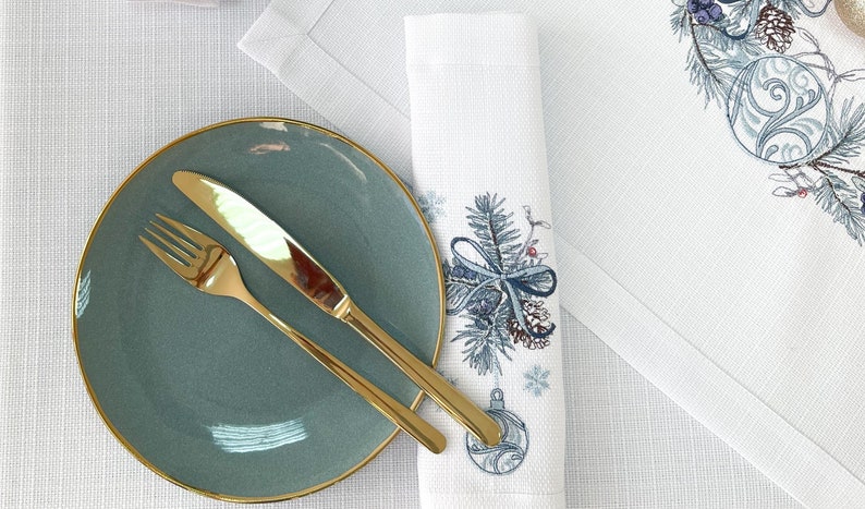 K&ouml;nnte beinhalten: Ein t&uuml;rkisfarbener Teller mit Goldrand, eine goldene Gabel und ein goldenes Messer liegen auf einer wei&szlig;en Tischdecke. Rechts befindet sich eine wei&szlig;e Serviette mit einem blau-silbernen Weihnachtsbaumdesign. Im Hintergrund ein wei&szlig;er Tischset mit passendem Design.