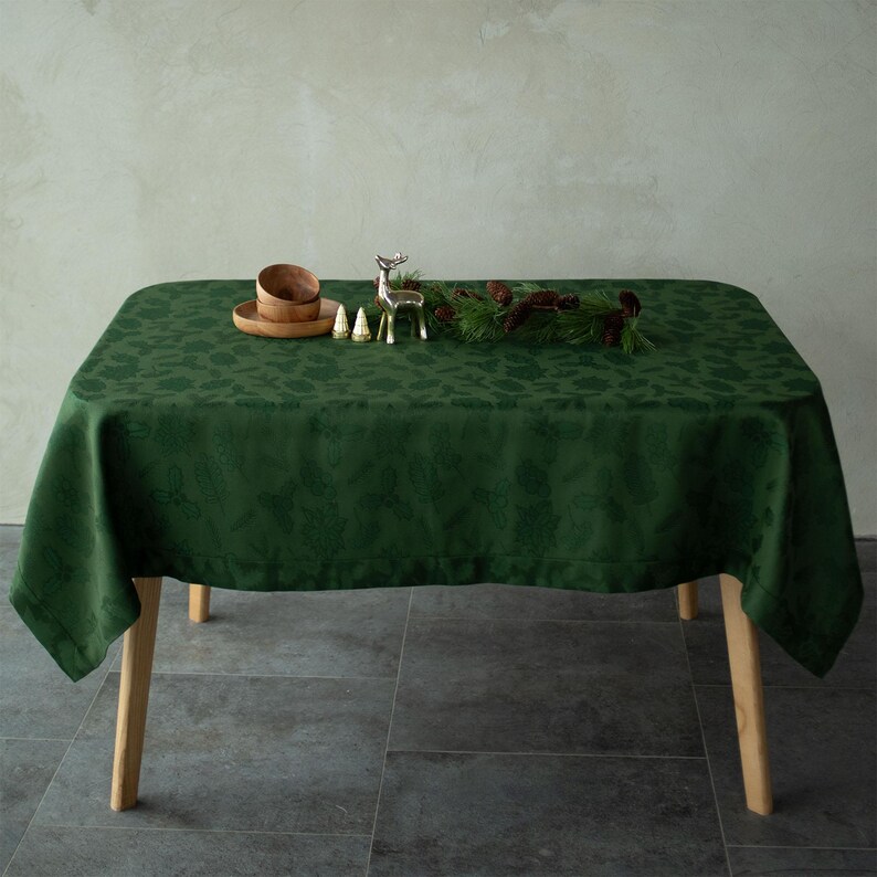Op de afbeelding: Een groene tafelkleed met een subtiel bloemmotief bedekt een tafel met houten poten. Decoratieve items zijn houten kommen, miniatuurbomen, een zilveren rendierfiguur en een dennen- en dennenappelkrans, wat een feestelijke setting cre&euml;ert.