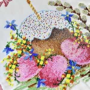 May include: Embroidered white fabric with a beaded Easter cake and lit candle, surrounded by beaded flowers and eggs. The top of the fabric has red text. The design is detailed and colourful.