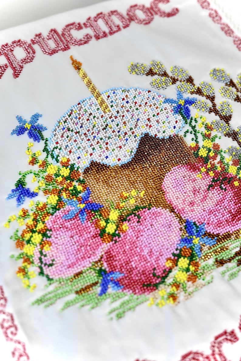 May include: Embroidered white fabric with a beaded Easter cake and lit candle, surrounded by beaded flowers and eggs. The top of the fabric has red text. The design is detailed and colourful.